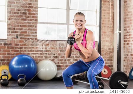 Adult female squatting with black kettlebell at chest in gym wearing pink sports bra blue leggings Adult female squatting with black kettlebell at chest in gym wearing pink sports bra blue leggings 141545603