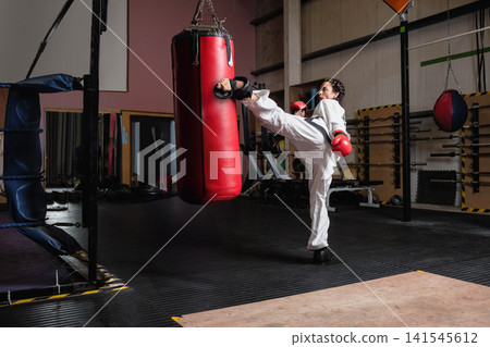 Male kicking heavy punching bag in gym while wearing gi, gloves, foot guards, copy space Male kicking heavy punching bag in gym while wearing gi, gloves, foot guards, copy space 141545612