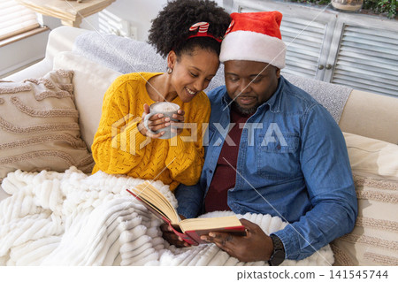 African American couple sitting on sofa at home, man wearing Santa hat, woman holding gray mug African American couple sitting on sofa at home, man wearing Santa hat, woman holding gray mug 141545744
