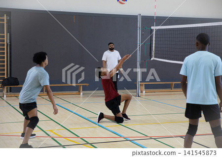 Male teenage players and coach training volleyball in gym with net, kneeling player reaching ball Male teenage players and coach training volleyball in gym with net, kneeling player reaching ball 141545873