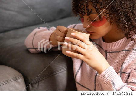 African American woman holding mug on grey sofa wearing pink grid sweater and red eye patches 141545881