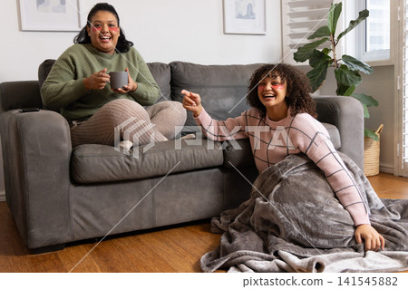 African American women in sweaters relaxing on gray sofa in living room, holding mug and pointing African American women in sweaters relaxing on gray sofa in living room, holding mug and pointing 141545882
