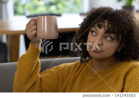 African American woman sitting on sofa holding beige mug near face wearing mustard-yellow sweater African American woman sitting on sofa holding beige mug near face wearing mustard-yellow sweater 141545883