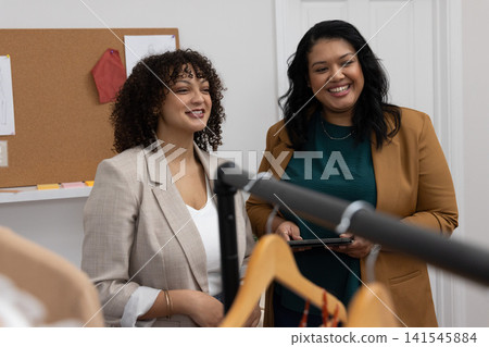 Diverse female colleagues discussing garments on rack with hangers in small showroom holding tablet Diverse female colleagues discussing garments on rack with hangers in small showroom holding tablet 141545884