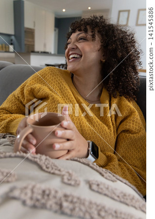 Woman in 30s reclining on gray sofa, wearing mustard sweater holding tan mug over beige throw Woman in 30s reclining on gray sofa, wearing mustard sweater holding tan mug over beige throw 141545886