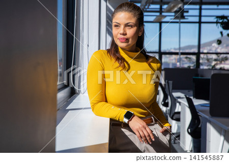 Adult woman leaning on window at office in mustard sweater and beige trousers, checking smartwatch Adult woman leaning on window at office in mustard sweater and beige trousers, checking smartwatch 141545887