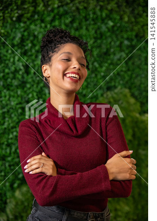 African American woman smiling and posing by green hedge wearing burgundy sweater and hoop earrings African American woman smiling and posing by green hedge wearing burgundy sweater and hoop earrings 141545888