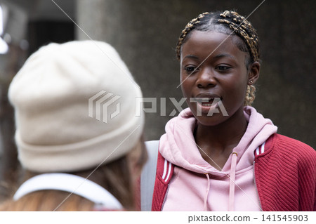 African American teenage girl talking on campus wearing red jacket backpack strap, friend in beanie African American teenage girl talking on campus wearing red jacket backpack strap, friend in beanie 141545903