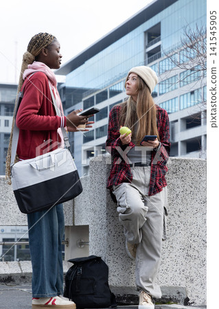Diverse female friends chatting on concrete ledge at plaza, holding smartphones and green apple 141545905