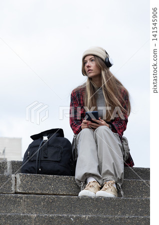 Woman sitting on concrete steps holding smartphone, wearing beanie headset plaid shirt and backpack 141545906