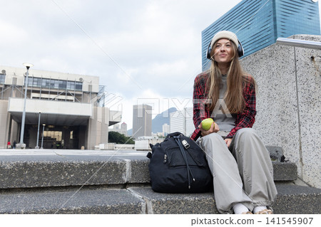 Woman sitting on steps at plaza holding green apple with black backpack, looking up, copy space Woman sitting on steps at plaza holding green apple with black backpack, looking up, copy space 141545907