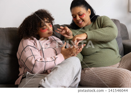 African American mom, teen daughter sitting on couch applying red under-eye patches with glass bowl African American mom, teen daughter sitting on couch applying red under-eye patches with glass bowl 141546044