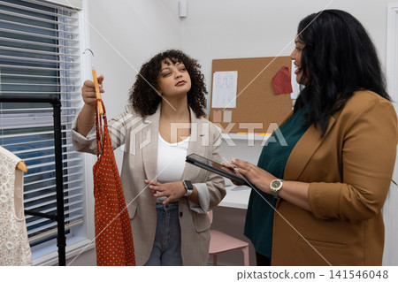 Diverse female client and stylist checking orange polka-dot dress on hanger, using tablet in shop Diverse female client and stylist checking orange polka-dot dress on hanger, using tablet in shop 141546048
