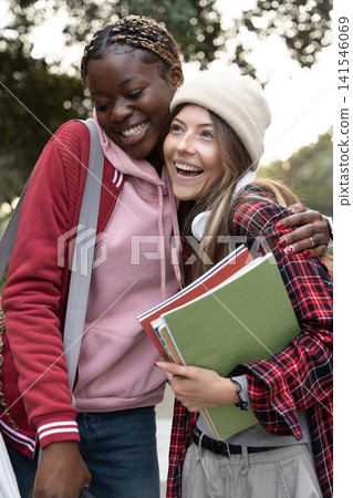 Diverse teen female students standing on tree-lined path, leaning and smiling, holding notebooks Diverse teen female students standing on tree-lined path, leaning and smiling, holding notebooks 141546069
