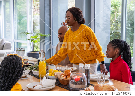 African American family sharing at dining table with woman in yellow sweater reaching orange juice African American family sharing at dining table with woman in yellow sweater reaching orange juice 141546075