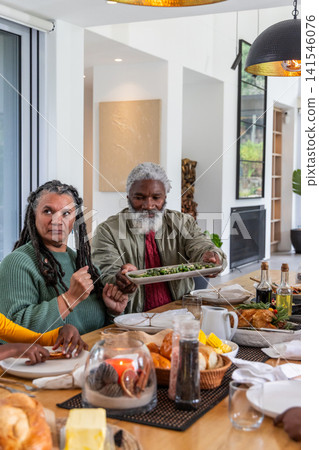 Senior couple passing green salad platter while woman in green sweater reaching across dining table Senior couple passing green salad platter while woman in green sweater reaching across dining table 141546076