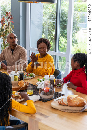 Diverse family sharing meal at home dining around wooden table with mom wearing mustard sweater Diverse family sharing meal at home dining around wooden table with mom wearing mustard sweater 141546080