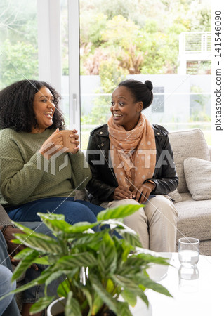 Two African American women sitting on sofa at home wearing sweater and leather jacket, holding mug Two African American women sitting on sofa at home wearing sweater and leather jacket, holding mug 141546090