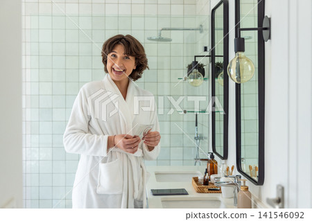 Middle-aged female wearing white terry robe standing at vanity holding floss near rain showerhead 141546092