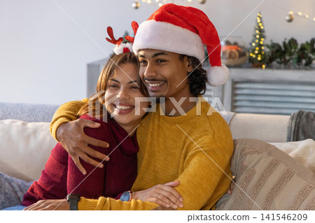 Diverse couple cuddling on sofa in living room wearing knit sweaters, Santa hat, with string lights Diverse couple cuddling on sofa in living room wearing knit sweaters, Santa hat, with string lights 141546209