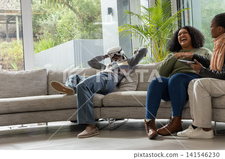 African American friends laughing, reclining on grey sofa in living room, one using VR headset African American friends laughing, reclining on grey sofa in living room, one using VR headset 141546230