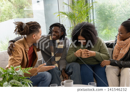 African American friends sitting closely on light-colored sofa in living room, holding ceramic mugs African American friends sitting closely on light-colored sofa in living room, holding ceramic mugs 141546232