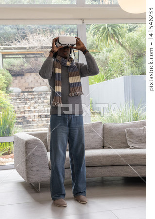 African American man standing in modern living room by large glass wall holding white VR headset African American man standing in modern living room by large glass wall holding white VR headset 141546233