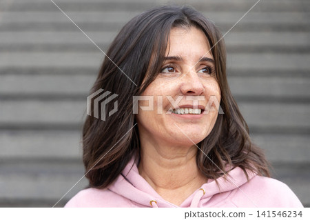 Woman smiling in light pink hoodie, looking slightly right on wide concrete steps Woman smiling in light pink hoodie, looking slightly right on wide concrete steps 141546234