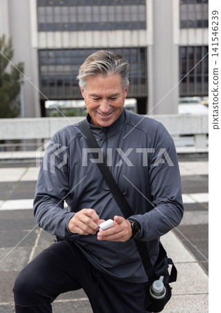 Middle-aged man leaning on plaza ledge opening earbud case in jacket with crossbody bag and bottle Middle-aged man leaning on plaza ledge opening earbud case in jacket with crossbody bag and bottle 141546239