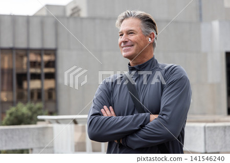Senior man standing near rooftop concrete railing, smiling, looking right, wearing earbuds Senior man standing near rooftop concrete railing, smiling, looking right, wearing earbuds 141546240