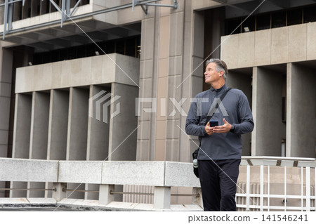 Middle-aged man checking smartphone on raised walkway by office, wearing crossbody bag, copy space 141546241