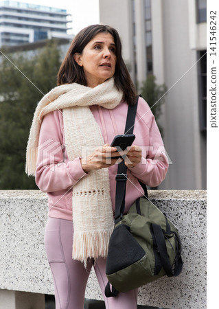 Woman standing outside by concrete wall holding phone with duffel wearing pink hoodie, cream scarf Woman standing outside by concrete wall holding phone with duffel wearing pink hoodie, cream scarf 141546242