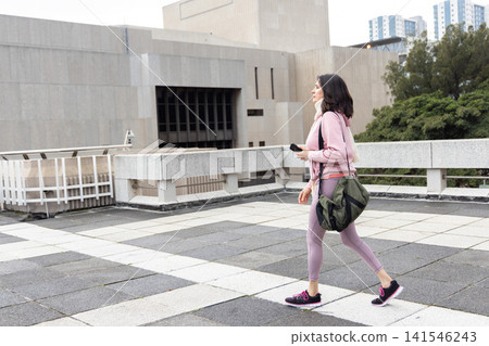 Asian woman walking on tiled plaza in pink hoodie, carrying green bag, holding phone, copy space Asian woman walking on tiled plaza in pink hoodie, carrying green bag, holding phone, copy space 141546243