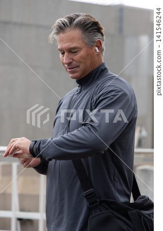Middle-aged man standing on elevated walkway checking watch wearing earbud dark jacket with duffel Middle-aged man standing on elevated walkway checking watch wearing earbud dark jacket with duffel 141546244