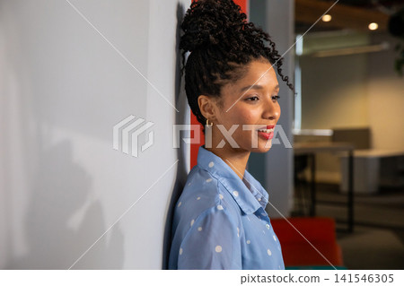 African American woman leaning on pale wall in office, wearing blue polka-dot blouse, copy space 141546305