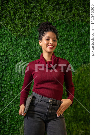 African American woman standing against plant wall, wearing maroon sweater and belt, holding tablet 141546309