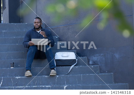 African American man sitting on steps using laptop near shoulder bag holding coffee cup, copy space African American man sitting on steps using laptop near shoulder bag holding coffee cup, copy space 141546318