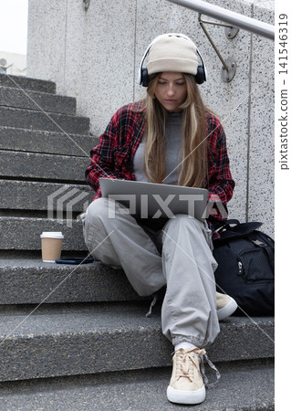 Woman in her 20s sitting on concrete steps at campus, using laptop wearing beanie and headphones Woman in her 20s sitting on concrete steps at campus, using laptop wearing beanie and headphones 141546319
