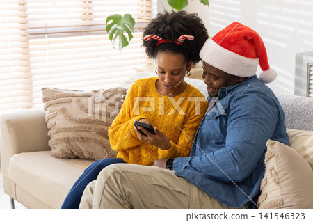 African American father and teen daughter sitting on sofa, sharing phone, Santa hat, candy headband African American father and teen daughter sitting on sofa, sharing phone, Santa hat, candy headband 141546323