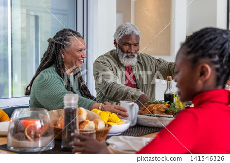 Senior African American couple smiling, passing roasted bird and bread at home dining table Senior African American couple smiling, passing roasted bird and bread at home dining table 141546326