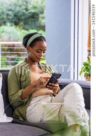 African American woman sitting on gray sofa by sliding door, holding smartphone wearing headband African American woman sitting on gray sofa by sliding door, holding smartphone wearing headband 141546334