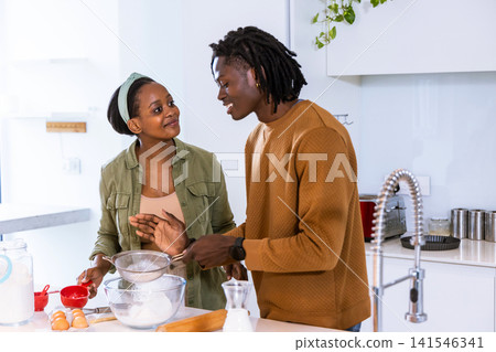 African American couple preparing baking at kitchen island with glass bowl, sifter, eggs, milk 141546341