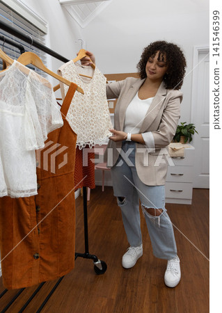 African American woman holding white lace top, inspecting rolling rack in fitting room in blazer African American woman holding white lace top, inspecting rolling rack in fitting room in blazer 141546399