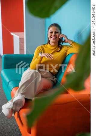 Woman sitting on teal orange sofa in lounge, in mustard top, holding smartphone, smartwatch visible Woman sitting on teal orange sofa in lounge, in mustard top, holding smartphone, smartwatch visible 141546408