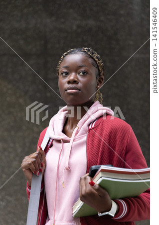 African American teen wearing pink hoodie and red jacket holding folders and bag strap at school 141546409