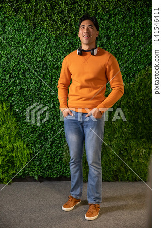 Asian male mid 20s standing on carpet before green foliage wall in orange sweatshirt and headphones Asian male mid 20s standing on carpet before green foliage wall in orange sweatshirt and headphones 141546411