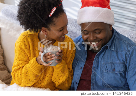 African American couple sitting on sofa at home holding frothy grey mug and wearing Santa hat African American couple sitting on sofa at home holding frothy grey mug and wearing Santa hat 141546425