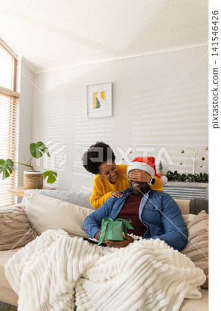 African American couple covering man's eyes on sofa, man wearing Santa hat holding green gift 141546426