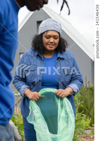 African American volunteers holding sheer green plastic bag in garden by grey home, with knit hat African American volunteers holding sheer green plastic bag in garden by grey home, with knit hat 141546438