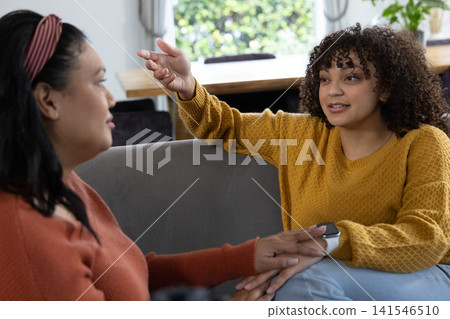 Two women sitting on grey sofa in living room, holding hands, talking by window with wristwatch 141546510